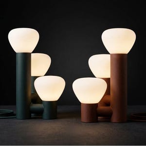 Peut inclure: Deux lampes de table à plusieurs niveaux, l'une vert foncé et l'autre brun terre cuite. Chaque lampe présente trois bases cylindriques de hauteurs variées, surmontées d'abat-jours en verre blanc arrondi diffusant une lumière douce.