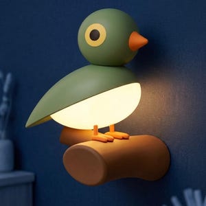 May include: A wall-mounted bird-shaped lamp featuring a smooth olive green head and wing, a bright orange beak, and a glowing white belly. The bird is perched on a brown cylindrical branch-like base that extends from the wall, providing illumination.
