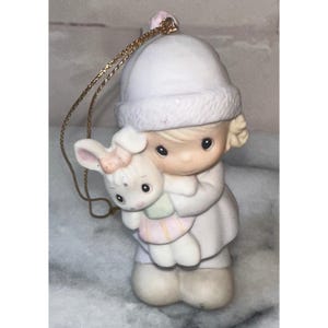 Könnte beinhalten: Eine matte Porzellanfigur als Anhänger, die ein Kind mit weißer Wintermütze und Mantel zeigt, das einen kleinen weißen Hasen mit rosa Schleife und pastellfarbenem Kleid hält. Eine goldene Kordel ist oben zur Aufhängung angebracht.