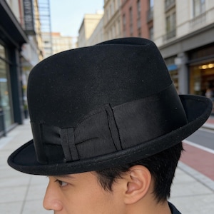 May include: A black felt fedora hat featuring a structured crown and a slightly curved brim. The hat is accented with a wide, matching black grosgrain ribbon band that includes a decorative bow detail on the side, encircling the base of the crown.