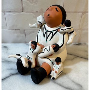 Pode incluir: Uma estatueta de cerâmica de contador de histórias com uma figura central grande de rosto terracota e cabelo preto, vestindo uma peça branca com detalhes pintados em preto e turquesa, rodeada por quatro figuras menores presas ao corpo.