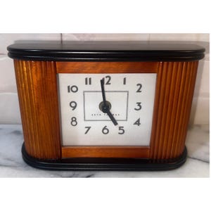 Puede incluir: Un reloj de sobremesa Seth Thomas con esfera blanca rectangular, números arábigos negros y manecillas negras. El reloj presenta un cuerpo de madera pulida en tono marrón medio con paneles laterales estriados y base negra.