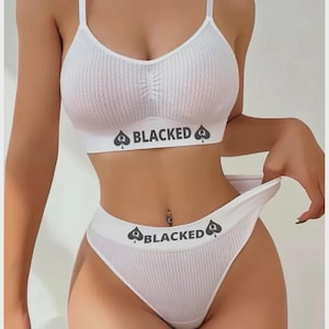 Puede incluir: Un conjunto de lencería de dos piezas en color blanco con textura acanalada, compuesto por un top corto y una braga de corte alto. Ambas piezas incluyen una banda elástica ancha con el texto BLACKED flanqueado por símbolos de picas con la letra Q.