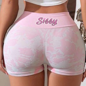 May include: High-waisted athletic shorts in a light pink and white tie-dye pattern. The waistband features the word Sissy printed in a stylized pink font. The shorts are made of a ribbed, seamless knit fabric with a fitted silhouette.