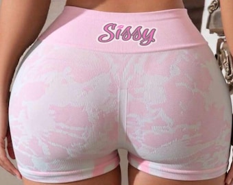 May include: High-waisted athletic shorts in a light pink and white tie-dye pattern. The waistband features the word Sissy printed in a stylized pink font. The shorts are made of a ribbed, seamless knit fabric with a fitted silhouette.