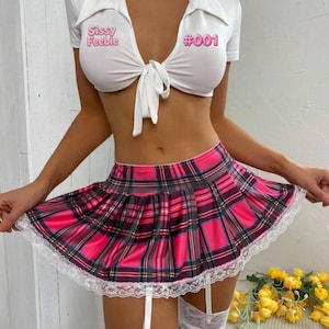 Può includere: Un top corto bianco a maniche corte con colletto e allacciatura frontale, con la scritta rosa Sissy Feebie e #001, abbinato a una minigonna a pieghe a quadri rosa, neri e blu con bordo in pizzo bianco e reggicalze bianchi integrati.