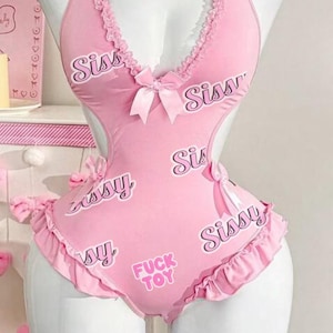 May include: A light pink bodysuit featuring a halter neckline with ruffled trim, a central bow, and cut-out sides. The fabric is printed with the words Sissy and FUCK TOY in a dark pink font. The garment includes additional small pink bows at the hips.