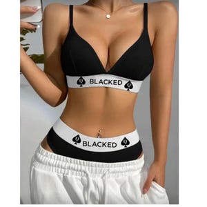 May include: A black two-piece lingerie set consisting of a bralette and matching briefs. Both pieces feature a wide white elastic waistband with the text BLACKED printed in black, flanked by two black spade symbols on either side of the word.