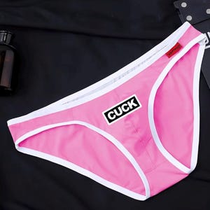 May include: Bright pink underwear featuring a white trim along the edges and a small red tag on the waistband. A rectangular black patch with the white text CUCK is centred on the front of the garment, which is made of a smooth fabric.