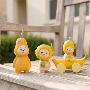 Könnte beinhalten: Drei geschnitzte Figuren in fruchtartigen Kostümen. Eine Figur ist birnenförmig und gelb, eine hat einen Zitronenkopf und trägt ein rosa Outfit, und eine hat einen Bananenkopf und sitzt auf einem gelben, bananenförmigen Wagen mit Rädern.
