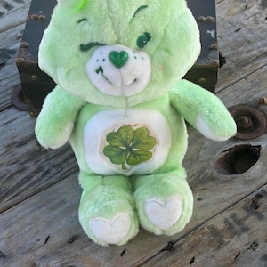 Peut inclure: Une peluche en forme d'ours vert clair avec un empiècement blanc sur le ventre orné d'un trèfle à quatre feuilles. L'ours a une expression clin d'œil, un nez vert en forme de cœur et des cœurs blancs sur la plante des pieds en peluche.