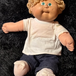 May include: A doll with light skin, bright green eyes, and curly blonde yarn hair. The doll wears a plain white short-sleeved t-shirt with a small tag on the left sleeve, dark blue denim pants, and white socks. The doll has a soft fabric body.