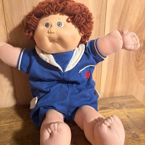 May include: A doll with curly reddish-brown yarn hair, a soft vinyl face, and a single visible tooth. The doll wears a blue sailor-style romper featuring a white collar, white pocket accents, and a small red embroidered anchor on the chest.