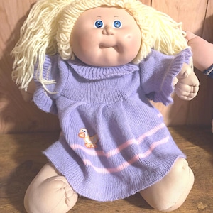 May include: A doll with light skin, blue eyes, and blonde yarn hair styled in pigtails. The doll wears a light purple knit dress featuring a ruffled collar, short sleeves, and a small embroidered duck graphic with two horizontal pink stripes.