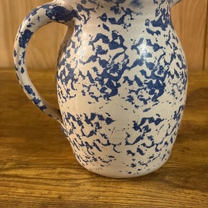 May include: A ceramic pitcher featuring a white base with a blue sponge-painted or speckled pattern. The vessel has a rounded body, a curved handle on the left side, and a small pouring spout at the top rim, showcasing a classic mottled design.