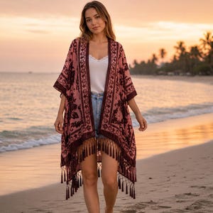 May include: A dusty rose kimono featuring dark burgundy velvet floral patterns, an open front, and a long fringe hem. The garment is worn over a white camisole and light blue denim shorts, showcasing a loose, flowing silhouette with wide sleeves.