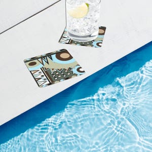 May include: Two square coasters featuring a geometric pattern in shades of light blue, brown, dark gray, and white. One coaster supports a clear glass filled with ice cubes and a lime wedge, while the other coaster sits empty nearby.