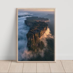 Può includere: Una stampa artistica da parete verticale incorniciata che mostra una vista aerea di una montagna dalla cima piatta, chiamata tepui, che si erge sopra uno strato denso di nuvole bianche. La parete rocciosa è illuminata da una calda luce solare dorata.