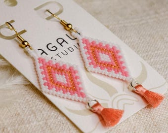 Pode incluir: Um par de brincos pendentes com um padrão de losango feito de missangas brancas, rosa claro e douradas. Cada brinco inclui uma pequena borla de cor coral na parte inferior e está preso a um gancho de metal de tom dourado.