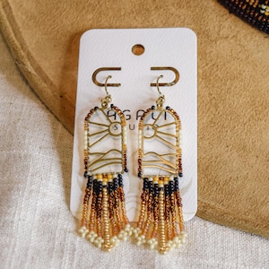May include: Dangle earrings featuring a gold-toned metal arch frame with a cutout landscape design of a sun and mountains. The lower portion consists of multi-coloured seed bead fringe in shades of black, brown, tan, and cream, attached to a white card.