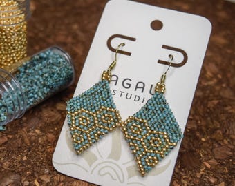 Pode incluir: Um par de brincos em forma de diamante feitos de pequenas contas turquesa e douradas, apresentados num cartão branco com a etiqueta AGAL STUDIO. Dois frascos de plástico transparente contêm contas douradas e turquesa.