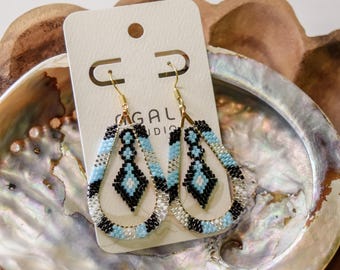 Pode incluir: Brincos em forma de gota feitos de pequenas contas num padrão geométrico preto, azul claro e branco. Os brincos apresentam ganchos de metal dourado e são exibidos num cartão branco retangular com o texto AGALI STUDIO.