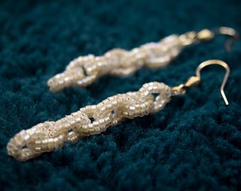 Pode incluir: Um par de brincos pendentes com um design de elos de corrente, feitos de pequenas contas iridescentes na cor branco sujo. Cada brinco está preso a um gancho de metal dourado, criando uma aparência texturizada e pendente com um brilho sutil.