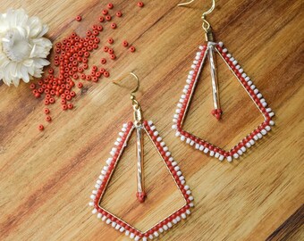May include: A pair of geometric dangle earrings featuring a gold-toned metal frame adorned with small red and white beads. Each earring has a central vertical bar and a hook-style closure. A small white flower and loose red beads are nearby.