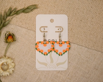 May include: A pair of heart-shaped beaded earrings featuring a white center surrounded by a border of small orange flowers and green leaves. The earrings are attached to a white rectangular display card with a sun graphic at the bottom.