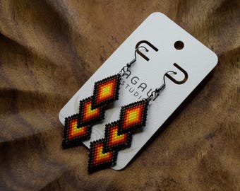 May include: A pair of dangle earrings featuring three stacked diamond-shaped beaded segments in a gradient of yellow, orange, and red, bordered by black beads. The earrings are attached to a white display card with the text AGALU STUDIO printed.