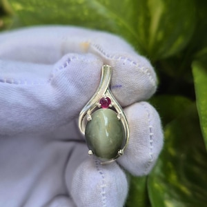 Op de afbeelding: Een zilveren hanger met een grote, ovale, grijsgroene cabochon edelsteen met een zichtbare chatoyante lichtband, geaccentueerd door een kleine, ronde, donkerroze edelsteen in een gepolijste metalen zetting met vier pootjes.