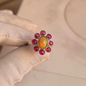 May include: A silver ring featuring a central oval-shaped iridescent opal cabochon surrounded by eight small, round, deep pink cabochon stones arranged in a floral pattern. The metal band is thin and polished, showcasing a bright, reflective finish.