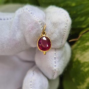 May include: A small, oval-shaped pendant featuring a faceted, deep pinkish-red gemstone set in a polished yellow gold bezel. The pendant includes a small gold jump ring at the top for attachment and a tiny gold bead detail at the bottom edge.