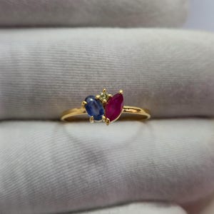 Puede incluir: Un anillo de oro con un grupo de tres piedras preciosas: un zafiro azul de talla ovalada, un rubí rojo de talla marquesa y un pequeño diamante redondo de color amarillo pálido engastado entre ellos en una banda de oro fina y pulida.