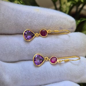Könnte beinhalten: Ein Paar goldfarbene Ohrhänger mit einem facettierten, birnenförmigen violetten Amethyst und einem kleineren runden rosa Edelstein. Die Ohrringe sind durch goldene Metallglieder verbunden und verfügen über einen einfachen Haken zur Befestigung.