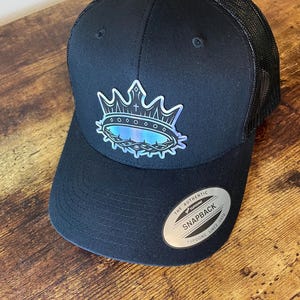 May include: A black snapback cap with a mesh back and a holographic crown graphic on the front. The crown design features a small cross and a thorn-like base. A silver circular sticker on the brim reads THE AUTHENTIC YUPOONG SNAPBACK.