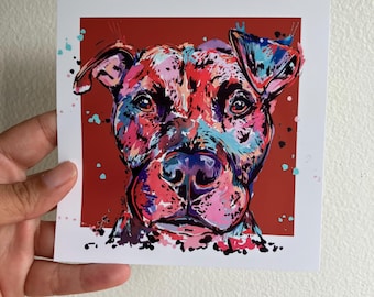 May include: A square art print featuring a vibrant, multi-colored portrait of a dog. The canine face is rendered in bold splashes of pink, blue, and purple against a solid dark red background, with paint splatter effects surrounding the head.