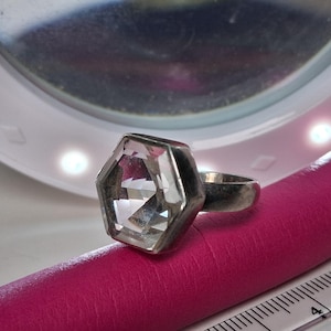 May include: A silver-toned ring featuring a large, clear, hexagonal-cut gemstone set in a metal bezel. The ring is positioned next to a white measuring tape showing a measurement of approximately 5 centimetres, highlighting the scale of the jewellery.