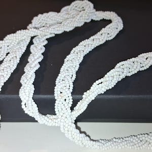 May include: A long, white beaded necklace featuring a complex braided design. The accessory is constructed from numerous tiny, uniform white beads woven together to create a textured, rope-like appearance that loops and drapes across the frame.