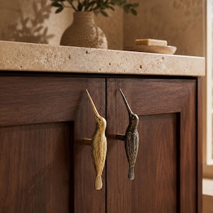 May include: Two cabinet pulls shaped like hummingbirds attached to dark wood cabinet doors. One pull is a polished gold tone, while the other is a dark, aged bronze finish. Both feature detailed wing and feather textures with long, pointed beaks.