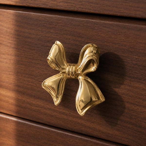 May include: A polished gold-toned metal cabinet knob shaped like a decorative bow with smooth, reflective surfaces and curved loops, mounted onto a dark brown wooden drawer front with a visible horizontal grain pattern and a clean finish.