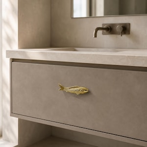 Puede incluir: Un tirador de mueble de metal dorado con forma de pez, con escamas y aletas detalladas, montado horizontalmente en un frente de cajón plano, mate y de color beige claro. El herraje presenta una superficie texturizada y una silueta realista.