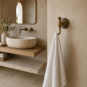 May include: A wall-mounted brass towel hook featuring a circular base with an intricate embossed border and a curved hook arm, holding a plain white fabric towel. The hardware has a warm metallic finish and is attached to a textured beige wall.