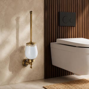 Puede incluir: Un escobillero de pared con acabado en latón antiguo, un recipiente de cerámica blanca con un borde dorado decorativo y un mango vertical largo. La placa base presenta un patrón en relieve ornamentado y se fija a la pared.
