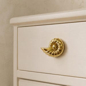 May include: A polished gold-toned metal cabinet knob shaped like a spiraled seashell. The knob is mounted on a smooth, off-white wooden drawer front. The seashell design features detailed, curved ridges radiating from a central circular point.