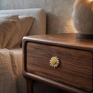 Könnte beinhalten: Ein Nachttisch aus dunklem Holz mit einer einzelnen Schublade und einem goldfarbenen, gänseblümchenförmigen Metallknopf. Die Schubladenfront zeigt eine ausgeprägte Holzmaserung, und der Knopf ist mittig platziert mit detaillierten Blütenblättern.