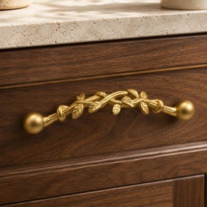May include: A gold-toned metal drawer pull featuring a central design of intertwined branches and small leaves, finished with smooth spherical knobs at each end. The handle is mounted horizontally on a dark brown wood cabinet drawer front.
