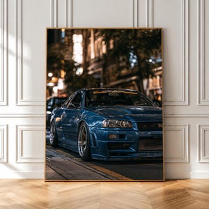 Könnte beinhalten: Ein gerahmter Wandkunstdruck, der einen dunkelblauen Nissan Skyline GT-R Sportwagen zeigt, der nachts auf einer Stadtstraße geparkt ist. Das Fahrzeug ist aus einer vorderen Dreiviertelansicht dargestellt und hebt den Metallic-Lack hervor.