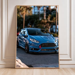 Op de afbeelding: Een ingelijste verticale wandkunstprint met een blauwe Ford Fiesta ST hatchback geparkeerd op een asfaltweg. De auto heeft een zwarte honingraatgrille, een rode sleepband en donkere meerspaaks wielen.