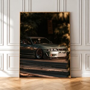 Peut inclure: Une impression d'art mural verticale encadrée représentant une voiture de sport Nissan Skyline GT-R R33 argentée garée sur une route goudronnée. Le véhicule est capturé en contre-plongée, mettant en valeur son pare-chocs avant et ses roues.